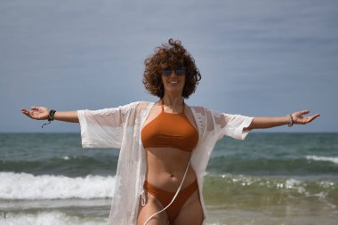 Attractive mature woman with curly hair, sunglasses and bikini, with open arms grateful and happy on the beach. Concept sea, sand, sun, beach, vacation, summer.