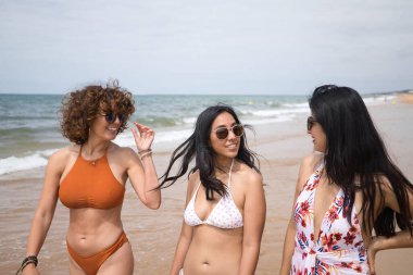 Attractive mature woman next to two young, pretty, brunette South American women, all wearing sunglasses and bikinis, walking and talking on the beach. Concept vacation, friends, multiethnic, summer.