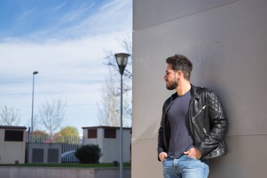 Handsome young man with beard, sunglasses, leather jacket and jeans, leaning against the wall of a building, hands in his pockets looking carefree. Concept beauty, fashion, trend, modern.