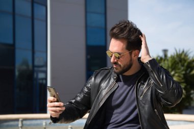 Handsome young man with beard, sunglasses, leather jacket and jeans, sitting, combing his hair while looking at the screen of his cell phone. Concept beauty, fashion, trend, app, smartphone, social.