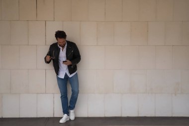 Sakallı, güneş gözlüklü, deri ceketli, beyaz gömlekli, kot pantolonlu, beyaz bir duvara yaslanmış, cep telefonuna danışan yakışıklı bir genç. Güzelliği, modayı, akıllı telefonu, uygulamayı, sosyal ağları kavra.