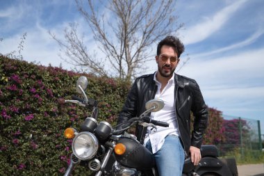 Sakallı, güneş gözlüklü, deri ceketli, beyaz gömlekli ve kot pantolonlu, büyük yer değiştiren motosikletli yakışıklı bir genç. Güzellik, moda, trend, motor, motosiklet, çıplak kavram.