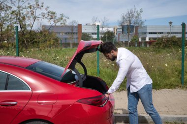 Sakallı, güneş gözlüklü, beyaz gömlekli, kot pantolonlu, kırmızı spor arabasının bagajını açan yakışıklı bir genç. Güzellik, moda, trend, lüks, motor, spor, başarılı, bagaj, bagaj.