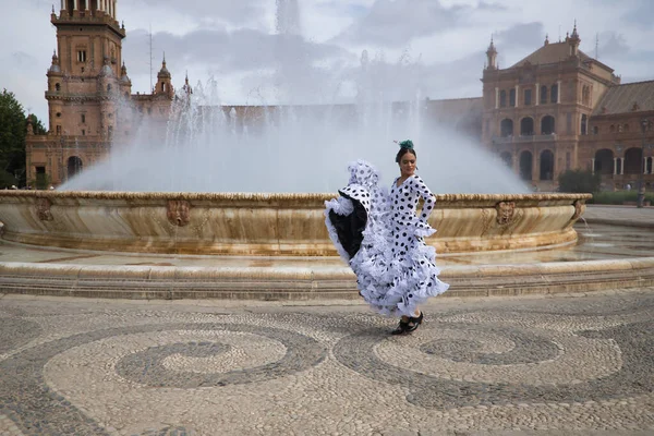 Young teenage woman in white dance suit with black polka dots and green carnations in her hair, dancing flamenco in front of a water fountain. Flamenco concept, dance, art, typical Spanish dance.