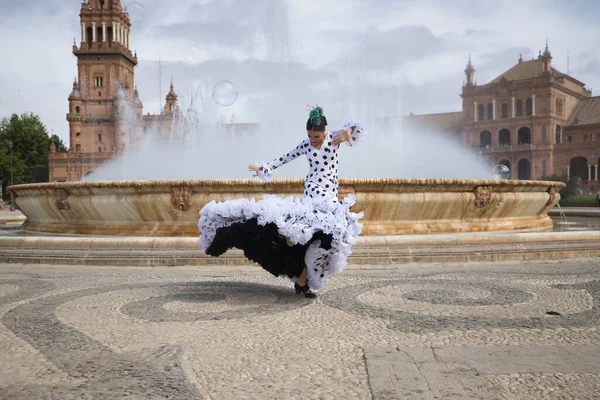 Young teenage woman in white dance suit with black polka dots and green carnations in her hair, dancing flamenco in front of a water fountain. Flamenco concept, dance, art, typical Spanish dance.