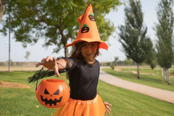 Girl in black shirt, witch hat and orange skirt with pumpkin on arm and hand trying to grab camera celebrating halloween. Autumn concept, trick or treat, party, pumpkin. Selective focus.