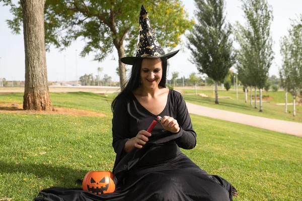 Woman in black witch suit and hat with a pumpkin, sitting on the grass while lighting a red candle, celebrating halloween. Autumn concept, trick or treat, party, pumpkin.