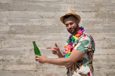 Young and handsome man, blue eyes, with beard, hat, Hawaiian shirt and flower necklace and pointing to a bottle of beer on a gray background. Concept vacation, party, travel, drink, copy space.