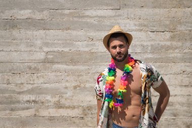 Young and handsome man, blue eyes, beard, hat, open Hawaiian shirt and flower necklace looking thoughtfully on a gray background. Concept holidays, party, trips.