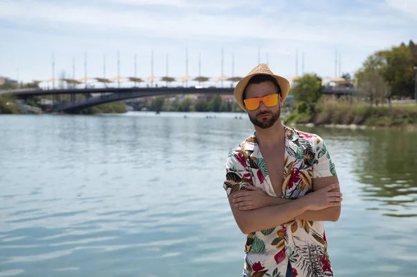 Young and handsome man, mirror sunglasses, beard, hat, Hawaiian shirt and jeans on a pier by the river, arms folded. Concept vacation, party, travel, cruises, sun.