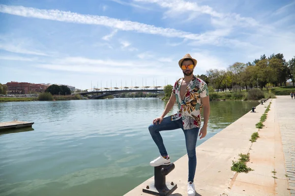 Young and handsome man, mirror sunglasses, beard, hat, Hawaiian shirt and jeans on a pier by the river. Concept vacation, party, travel, cruises, sun.