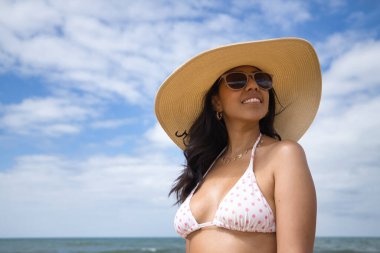 South American woman, young and beautiful, brunette with sunglasses, hat and bikini posing happy and smiling on the beach. Concept sea, sand, sun, beach, vacation, travel, summer.