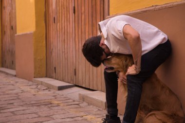 Young Hispanic man with beard, sunglasses and white shirt, leaning against a wall hugging his dog between his legs. Concept animals, dogs, love, pets, golden.