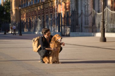 Young Hispanic man with beard, sunglasses, black shirt and backpack, crouched hugging his dog in the city. Concept animals, dogs, love, pets, golden.