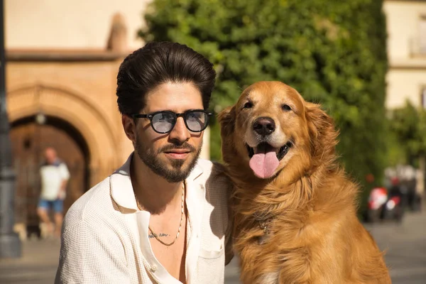 Portrait of young Hispanic man with beard, sunglasses and white shirt, sitting on a bench next to his precious dog. Concept animals, dogs, love, pets, golden.