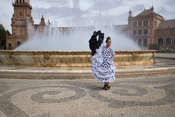 Young teenage woman in white dance suit with black polka dots and green carnations in her hair, dancing flamenco in front of a water fountain. Flamenco concept, dance, art, typical Spanish dance.