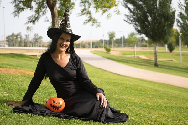 Woman in black witch suit and hat with a pumpkin, sitting on the grass in an outdoor park celebrating halloween. Autumn concept, trick or treat, party, pumpkin.