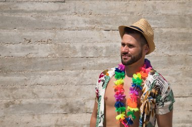 Young and handsome man, blue eyes, perfect smile, with beard, hat, open Hawaiian shirt and flower necklace looking thoughtfully on a gray background. Concept holidays, party, trips.