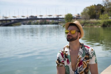 Young and handsome man, mirror sunglasses, beard, hat and Hawaiian shirt, sitting on a pier by the river. Concept vacation, party, travel, cruises, sun.