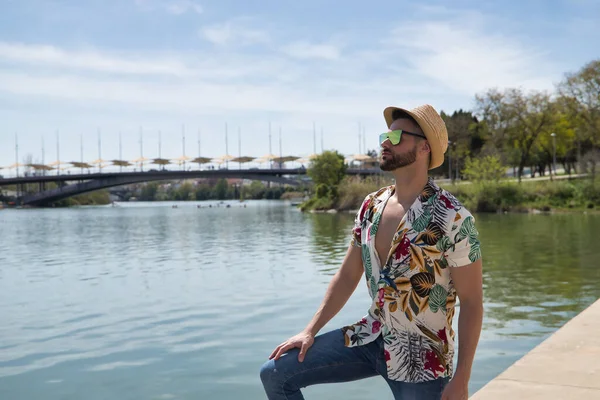 Young and handsome man, mirror sunglasses, beard, hat, Hawaiian shirt and jeans on a pier by the river. Concept vacation, party, travel, cruises, sun.
