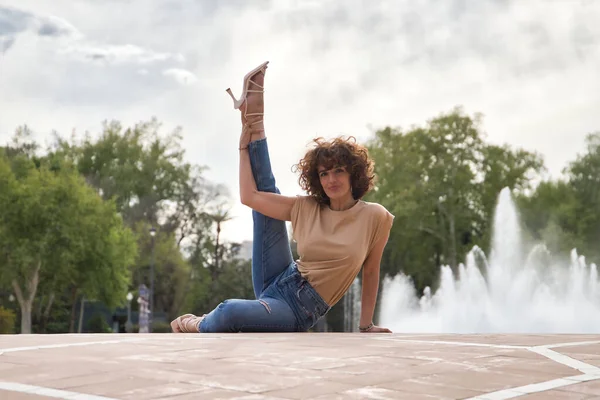 Mature and attractive woman, with curly brown hair, brown shirt, jeans and heeled shoes, sitting on the ground with one leg raised to the sky. Concept dance, trend, fashion, heels.