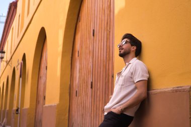 Young Hispanic man with beard and sunglasses leaning against a wall looking at the sky with distracted and thoughtful attitude. Concept thoughts, distraction, problems, anguish, depression.