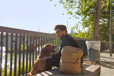 Young Hispanic man with beard, sunglasses, black shirt and backpack, sitting on a bench petting his dog under the sun rays. Concept animals, dogs, love, pets, golden.