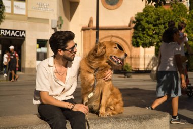 Young Hispanic man with beard, sunglasses and white shirt, sitting on a bench hugging and petting his dog. Concept animals, dogs, love, pets, golden.