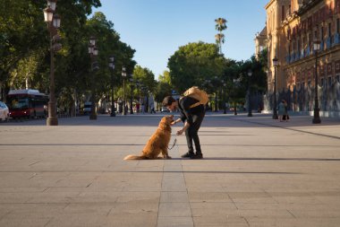 Young Hispanic man with a beard, sunglasses, black shirt and backpack, crouched down taking the leg of his dog in the city. Concept animals, dogs, love, pets, golden.