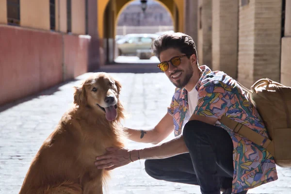 Portrait of young Hispanic man with beard and sunglasses crouching with his dog looking at camera on a sunny street at sunset. Concept animals, dogs, love, pets.
