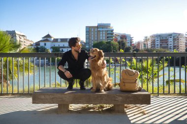 A young Hispanic man with a beard, sunglasses, black shirt and backpack, climbed on a bench with his dog while petting him under the sun's rays. Concept animals, dogs, love, pets, golden.