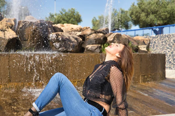 Attractive mature woman, wearing transparent black shirt and jeans, sitting next to a fountain, posing in sensual and provocative attitude. Concept maturity, beauty, fashion, seduction, provocation.