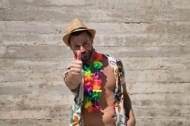 Young and handsome man, blue eyes, with beard, hat, open Hawaiian shirt and flower necklace and making ok sign with thumb on a gray background. Concept holidays, party, trips. Selective focus.