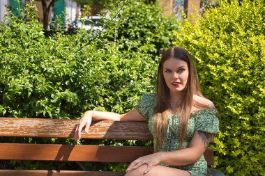 Genç, güzel bir kadın, sarışın, mavi gözlü, çiçekli yeşil bir takım elbise giyen, yeşil bitkilerden oluşan bir arka planda, ahşap bir bankta oturan. Güzelliği, modayı, eğilimi kavra.
