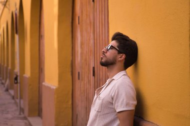 Young Hispanic man with beard and sunglasses leaning against a wall looking at the sky with distracted and thoughtful attitude. Concept thoughts, distraction, problems, anguish, depression.