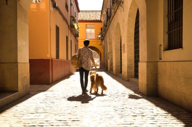 Young Hispanic man on his back with his dog holding the leash walking on a sunny street at sunset. Concept animals, dogs, love, pets, golden.