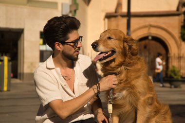 Young Hispanic man with beard, sunglasses and white shirt, sitting on a bench hugging and petting his dog. Concept animals, dogs, love, pets, golden.