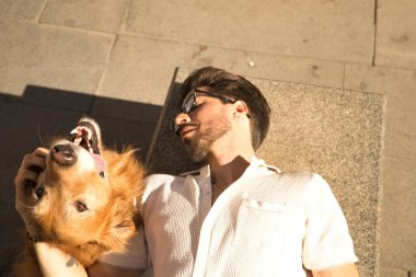 Young Hispanic man with beard, sunglasses and white shirt, lying on a bench petting his precious dog very happy. Concept animals, dogs, love, pets, golden.