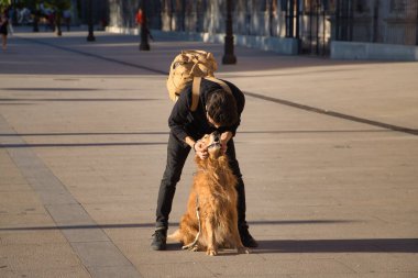 Young Hispanic man with beard, sunglasses, black shirt and backpack, crouched hugging his dog in the city. Concept animals, dogs, love, pets, golden.