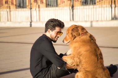 Sakallı, güneş gözlüklü, siyah gömlekli, yerde köpeğiyle oturan çok sevecen ve suç ortağı bir genç. Hayvanlar, köpekler, aşk, evcil hayvanlar, altın.