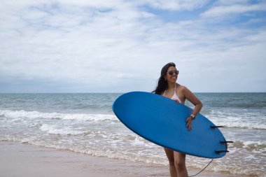 South American woman, young and beautiful, brunette with sunglasses and bikini, coming out of the water holding a blue surfboard. Concept sea, sand, sun, beach, vacation, surf, summer.