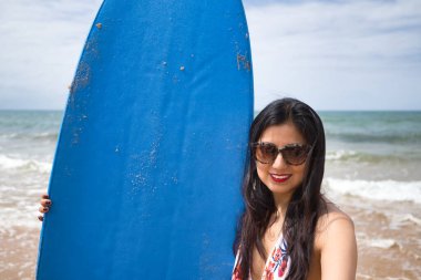 Güney Amerikalı bir kadın, genç ve güzel, güneş gözlüklü ve mayolu esmer, mavi bir sörf tahtası tutarken selfie çekiyor. Deniz, kum, güneş, sahil, tatil, sörf, yaz.