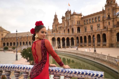 Kırmızı dans kıyafeti giymiş genç bir kadının portresi. Saçında kırmızı karanfillerle flamenko yapıyor. Korkuluklara yaslanmış pozlar veriyor. Flamenko konsepti, dans, sanat, tipik İspanyol dansı.