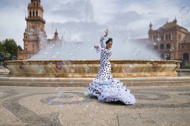 Siyah benekli beyaz takım elbiseli genç bir kadın çeşmenin önünde flamenko dansı yapıyor ve sabun köpükleriyle çevrili. Flamenko konsepti, dans, sanat, tipik İspanyol dansı, kabarcıklar.