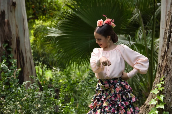 Young teenage woman in pink shirt, black skirt with flowers and pink carnations in her hair, dancing flamenco surrounded by greenery. Flamenco concept, dance, art, typical Spanish dance.