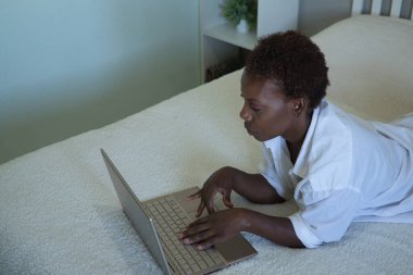 Beyaz gömlekli Afro-Amerikalı bir kadın ve yatakta dizüstü bilgisayarıyla çalışıyor. Telesekreter, internet, evden, yatak odasından çalışma kavramı.