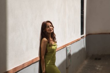 Dar yeşil takım elbiseli, uzun kahverengi saçlı ve tarçın tenli güzel bir kadın Akdeniz 'de dar bir caddenin beyaz duvarına yaslanıyor. Güzellik, moda, seyahat, tatil kavramları.