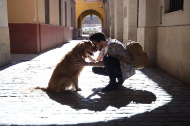Güneş batarken güneşli bir sokakta köpeğiyle çömelmiş sakallı ve güneş gözlüklü genç İspanyol adam gölgelerini yere düşürüyor. Hayvanlar, köpekler, aşk, hayvanlar.