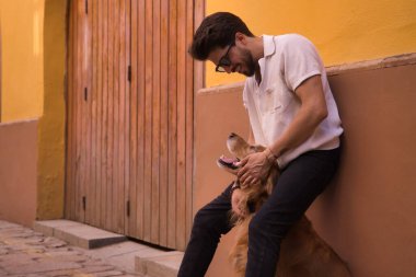Young Hispanic man with beard, sunglasses and white shirt, leaning against a wall hugging his dog between his legs. Concept animals, dogs, love, pets, golden.