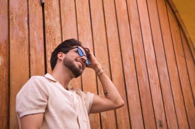 Young Hispanic man with beard, sunglasses and white shirt, combing his hair with his hand with a wall of wooden slats in the background. Concept model, hairstyle, hairdresser.